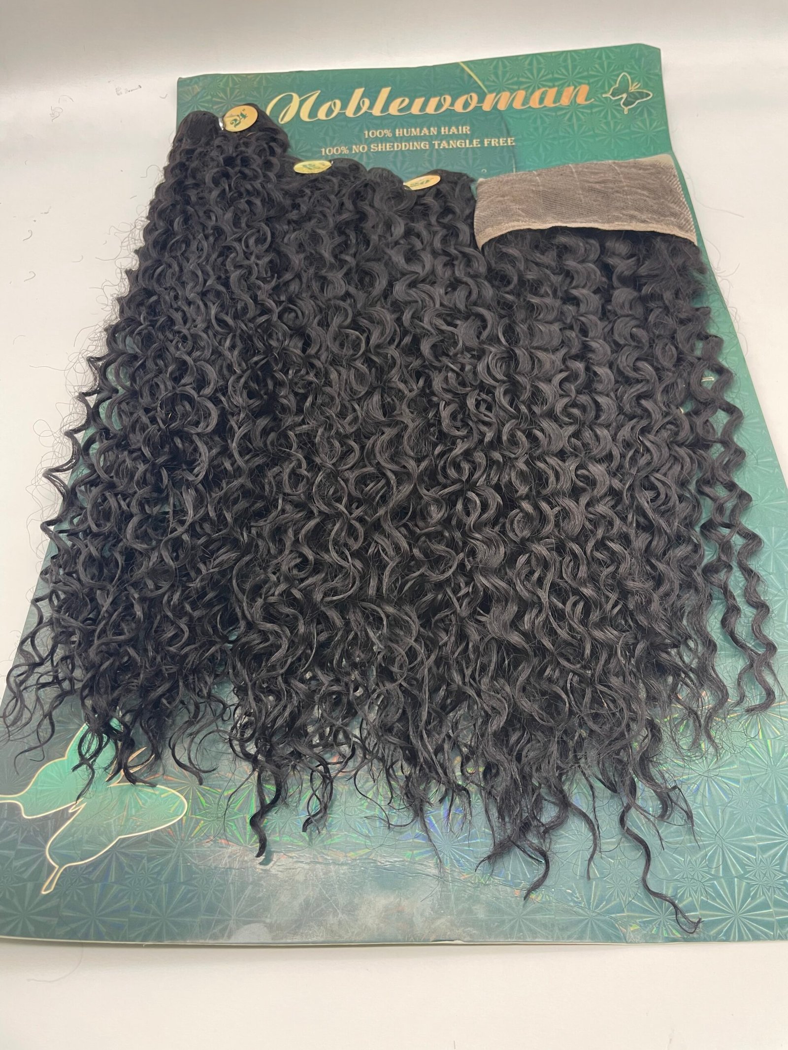 Bundles of tight black curly hair extensions laid across a green Noblewoman packaging card, labeled 100% human hair and no shedding tangle-free.