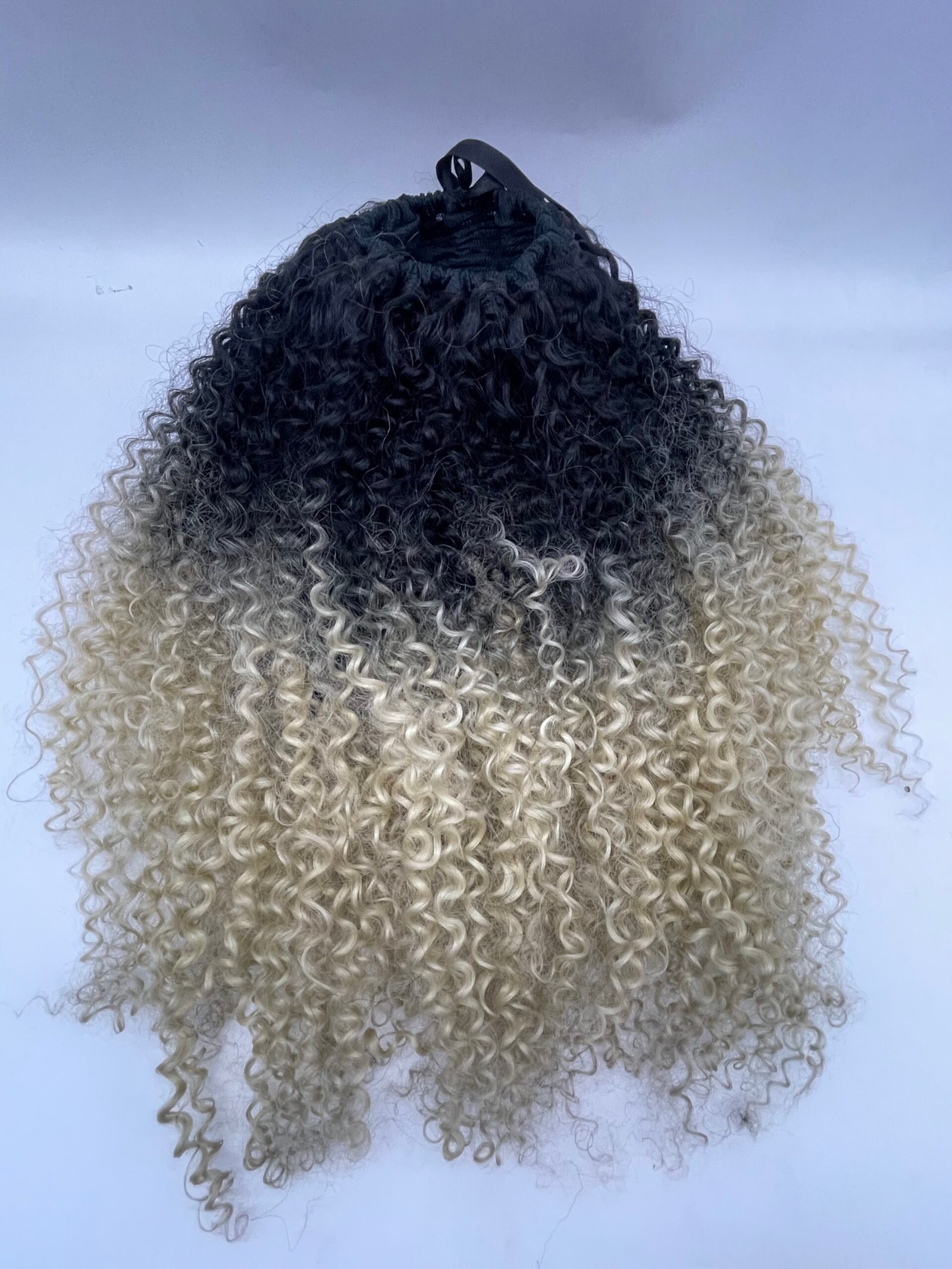 Back view of a person with long, tight curly hair transitioning from dark roots to blonde ends, held up with a black hair tie under a pale sky backdrop