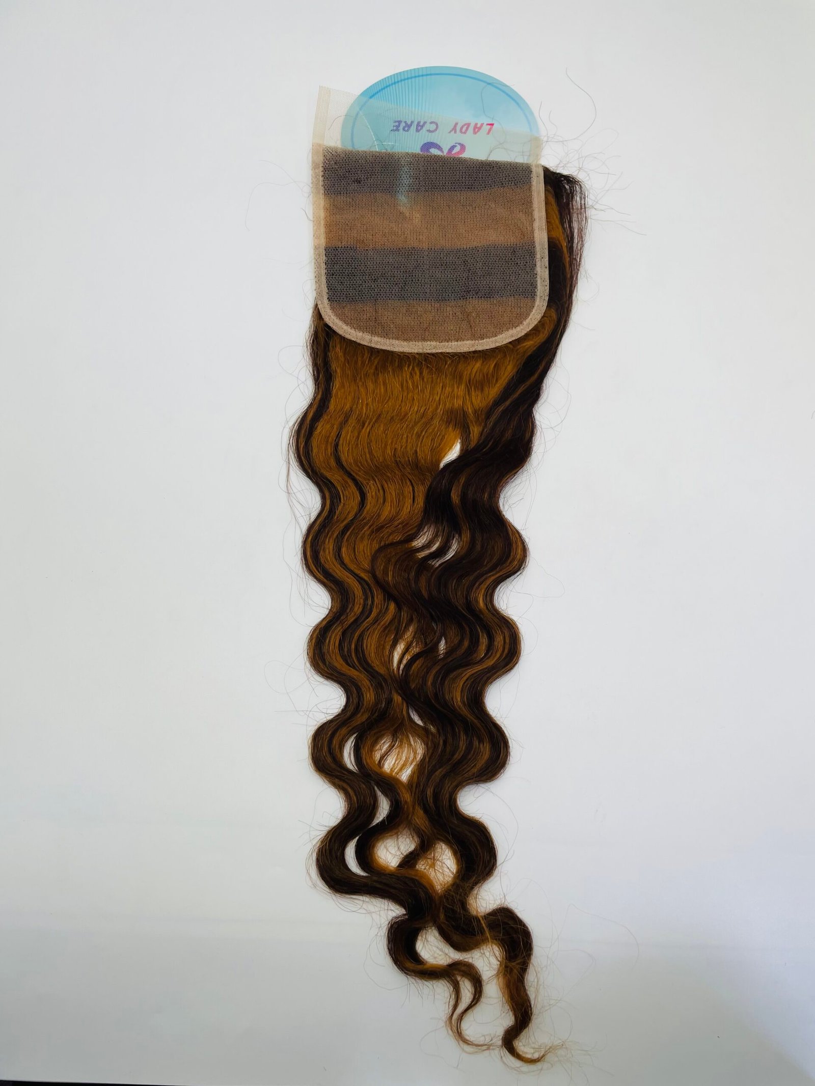 Wavy brown and black hair extension secured in a beige mesh cap with a blue disk behind it on a white background