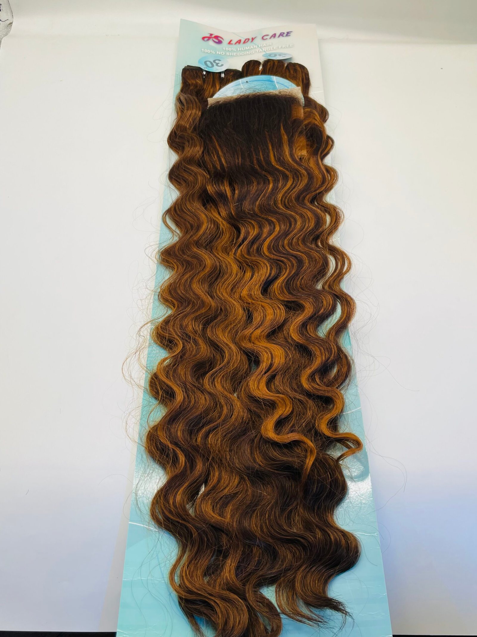 Copper-brown, wavy hair extensions mounted on a light blue backing with product packaging visible at the top.