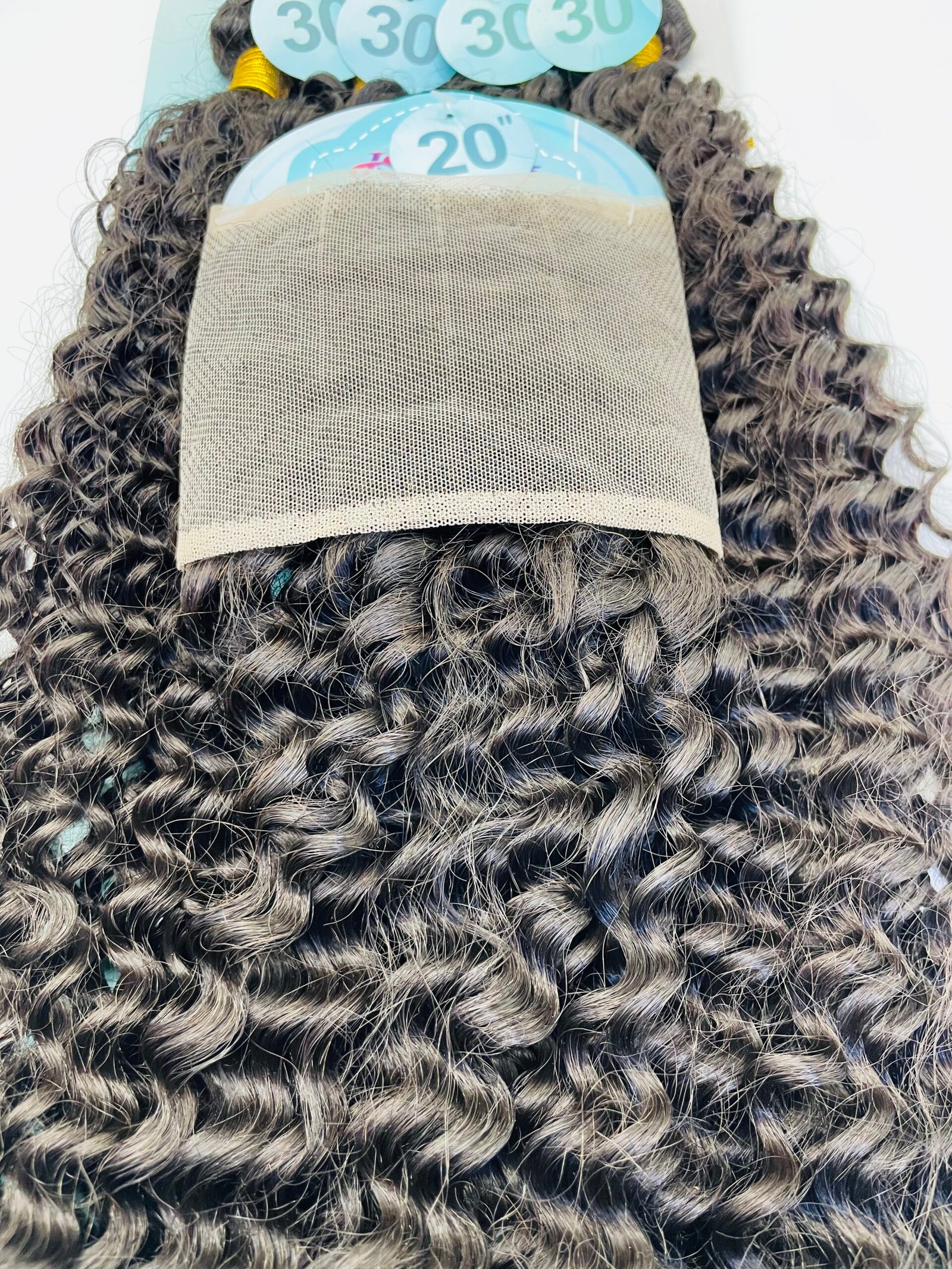 Close-up of a dark curly wig with a beige mesh wig cap draped over it; blue measurement discs (20") and (30") are visible behind the cap.