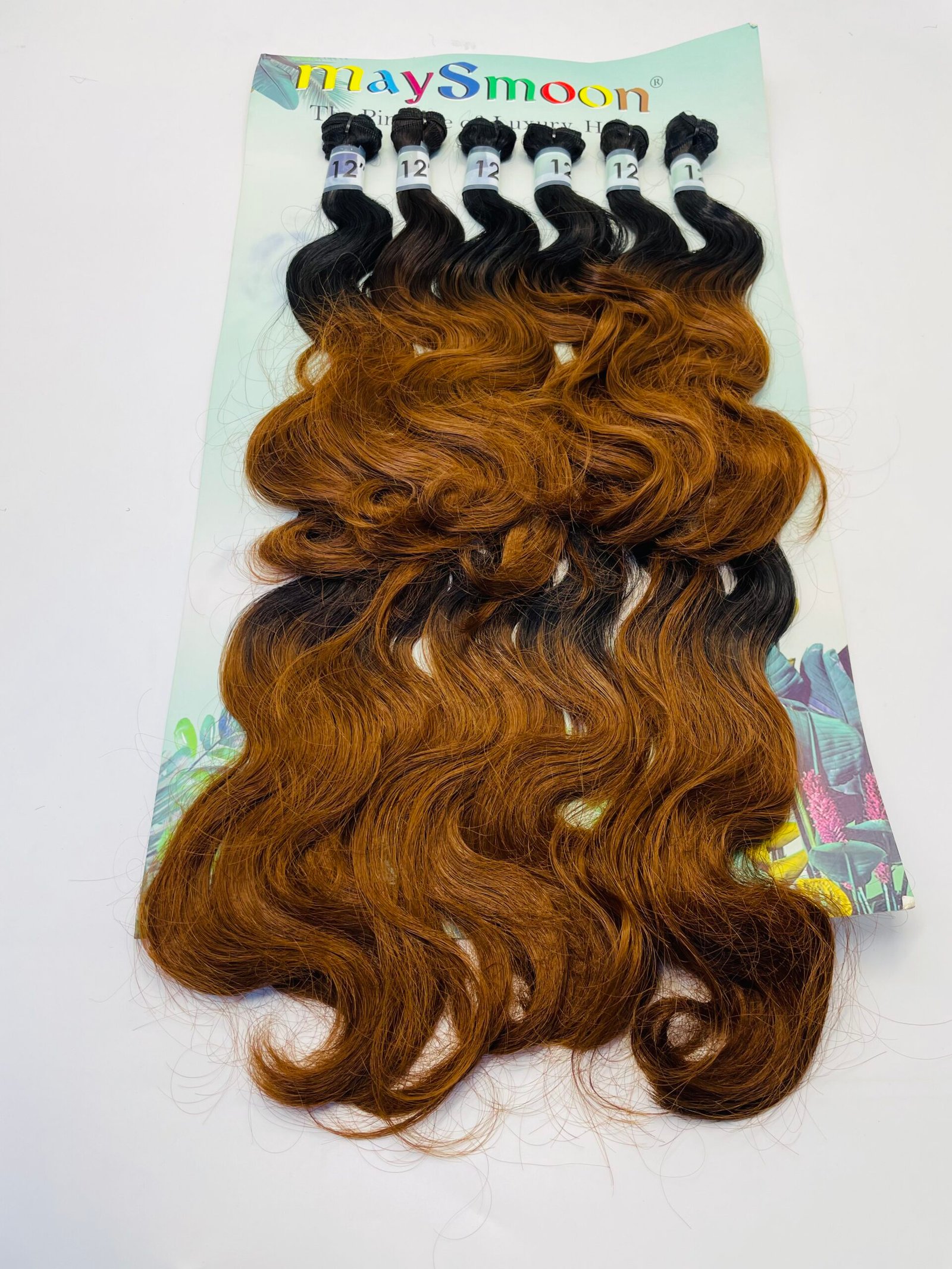 Six bundles of wavy hair extensions on a pastel display card, fading from black at the roots to copper brown at the ends.