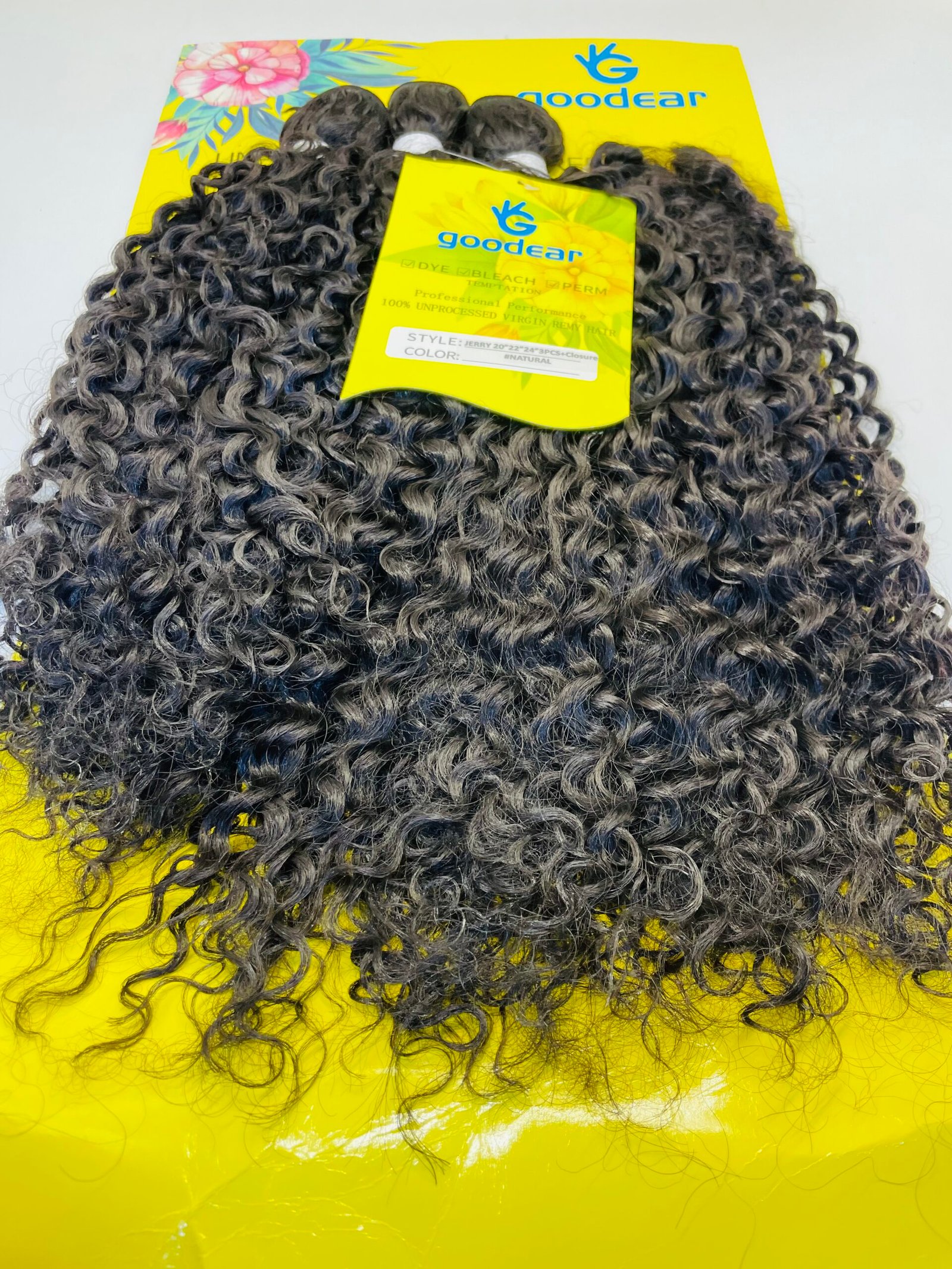 Dark curly hair extensions laid over a bright yellow packaging labeled 'goodear' with tropical flower graphics nearby.