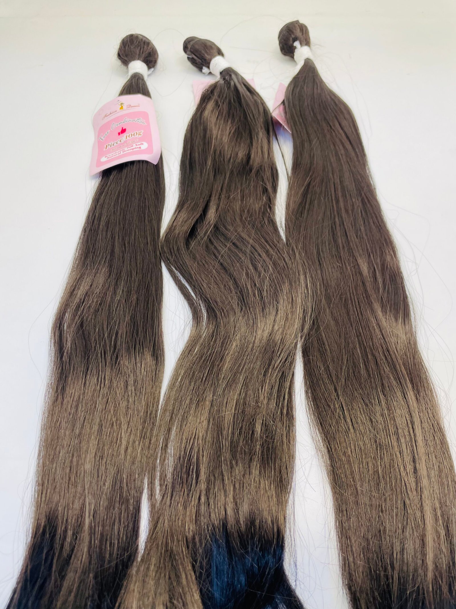 Three long brown hair extensions tied with white bands on a white surface, left one has pink tag attached in tag line style