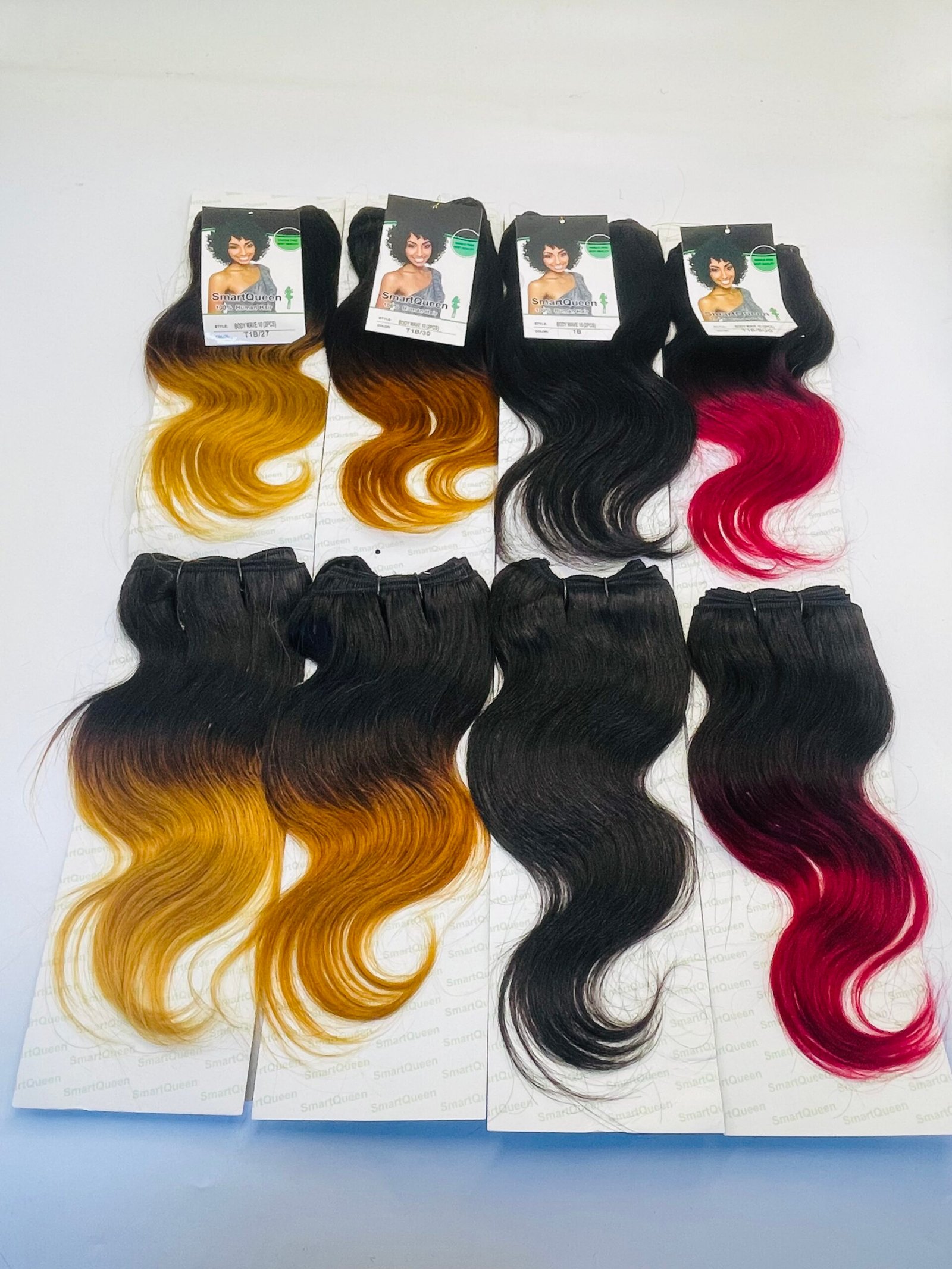 Four hair-extension bundles on white cards, each with wavy ombre color: yellow-to-blonde, orange-to-brown, solid black, and pink-to-red, with SmartQueen tags attached.