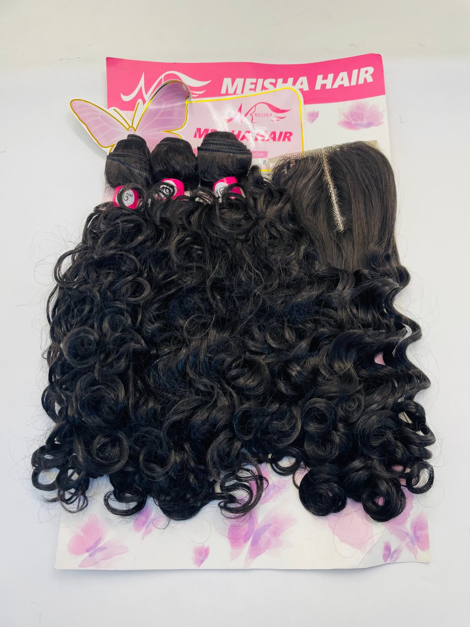 Curly black hair extensions laid over a pink Meisha Hair display card on a flower-patterned surface, viewed from above, showing texture and curl definition.