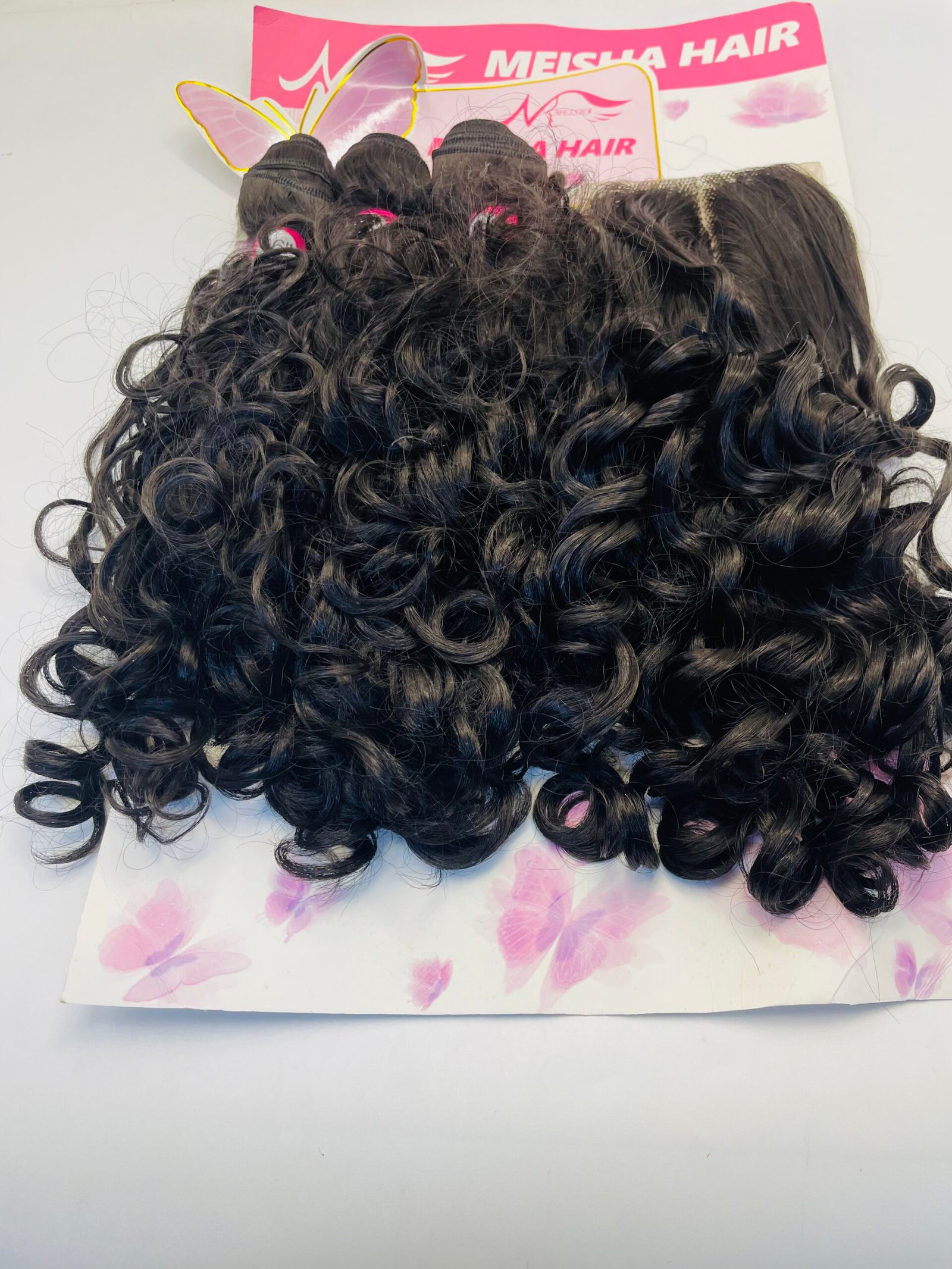 Close-up of glossy black tight curls with pink hair ties, set against a pink Meisha Hair sign backdrop.