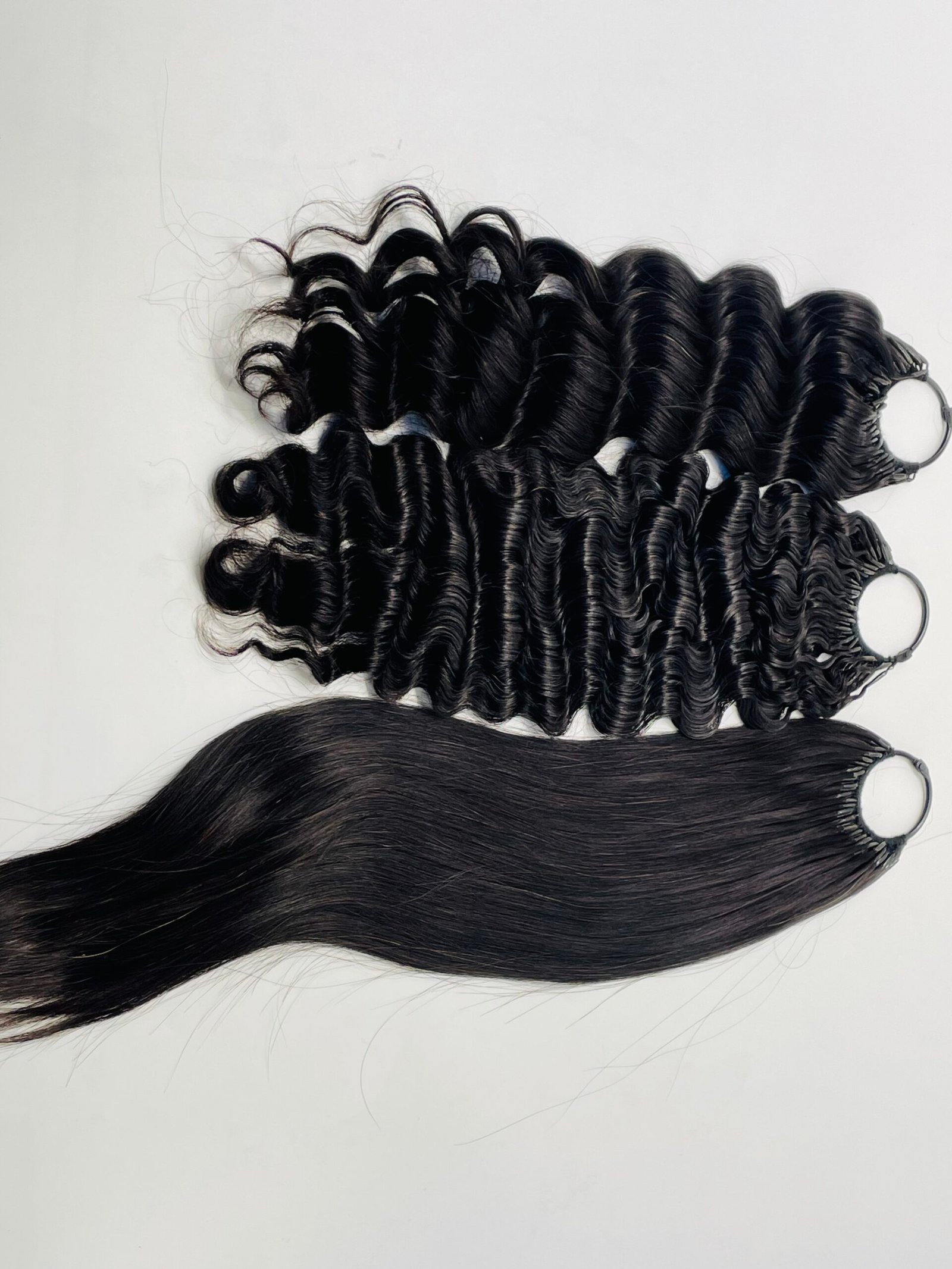 Bundles of glossy black curly hair extensions with metal rings at the top, laid on a light background.