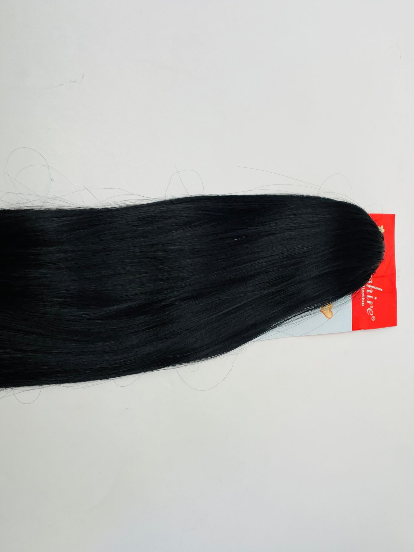 Bundle of long, straight black hair extensions laid across a white surface with a red packaging in the corner.
