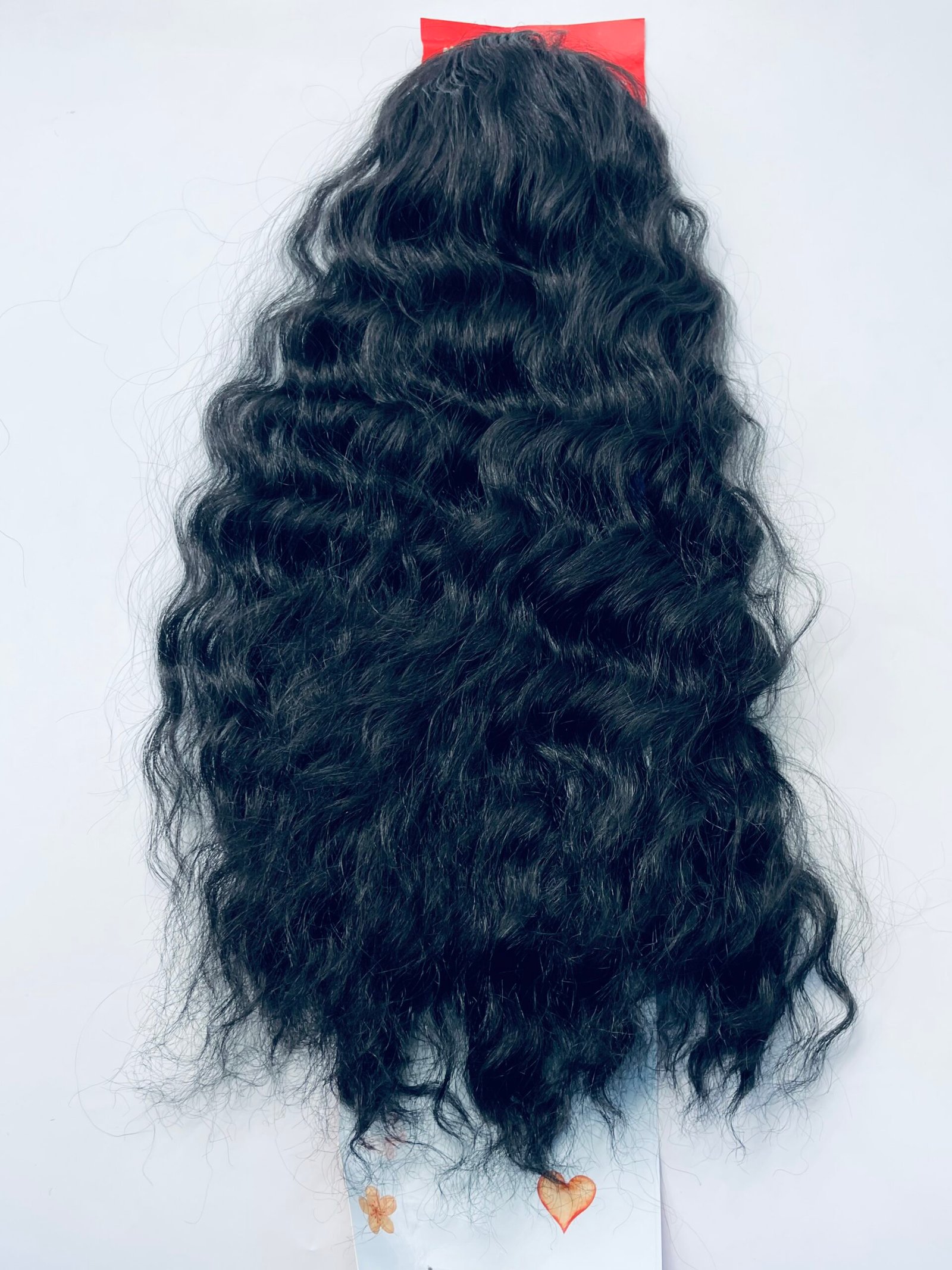 Back view of a person with long, wavy black hair; a small red square sits on top and a light blue strip with orange hearts hangs at the bottom.