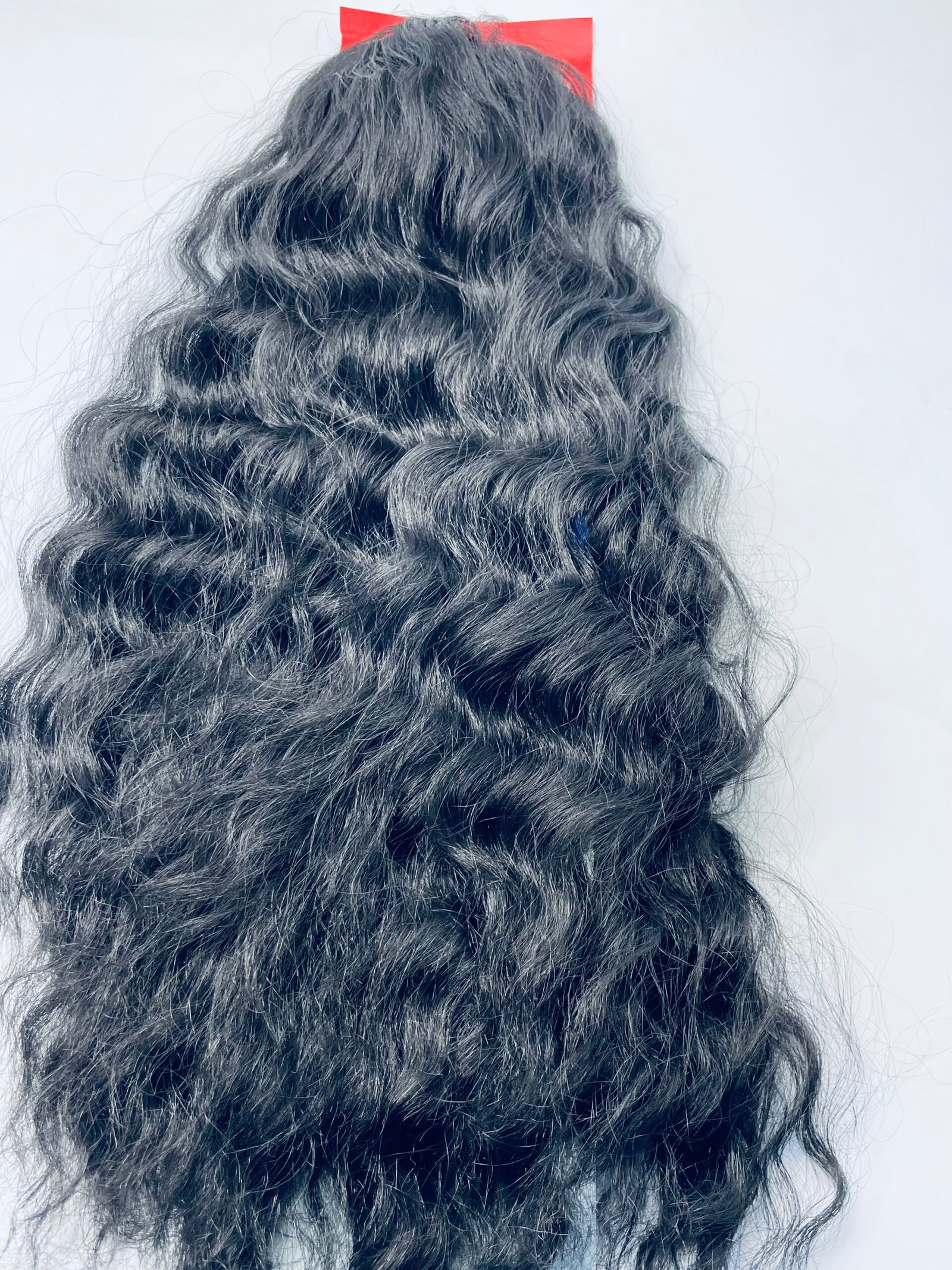 Back view of a person with long, curly black hair and a small red hair clip at the crown against a light background.