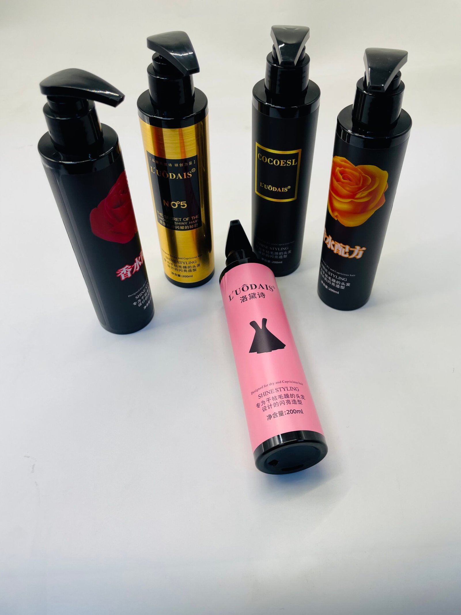 Group of black pump bottles for hair styling arranged on a white surface, with a pink bottle lying in front, highlighting branding and rose imagery on the labels.