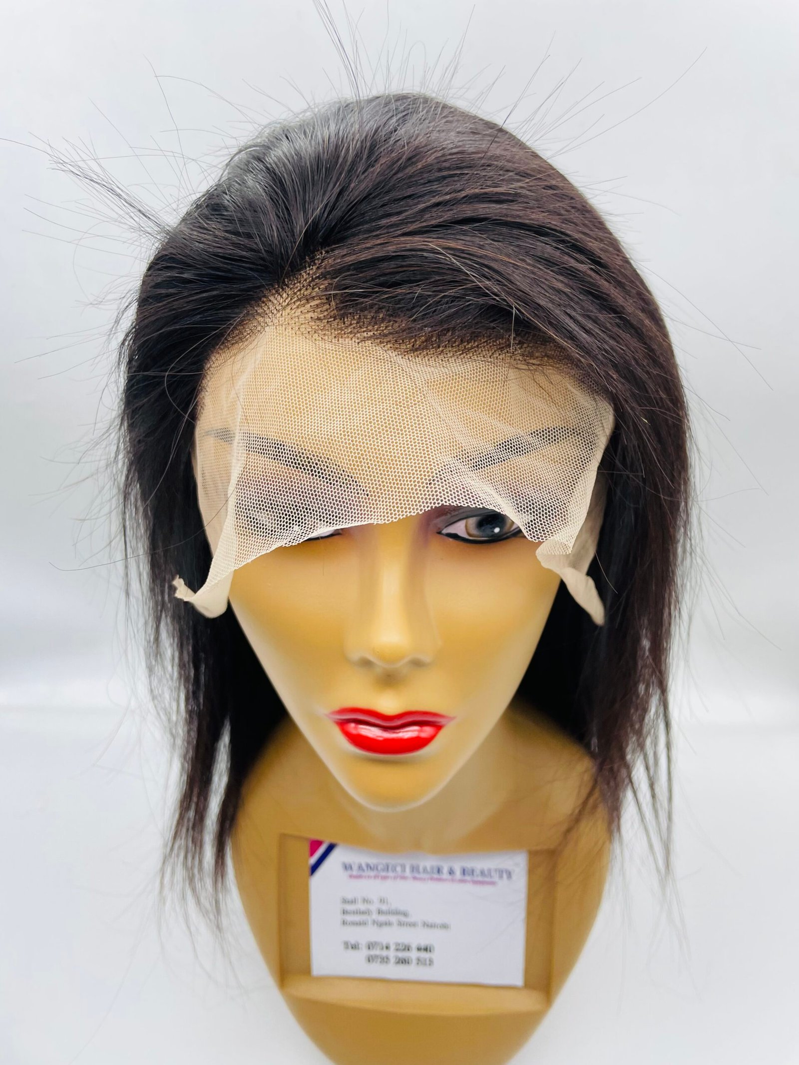 Mannequin head wearing a dark wig with a light mesh veil draped across the forehead and eyes; product label hanging at the neck.